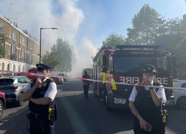 Police officers wearing masks guarding a road block in Maida Vale near substation fire on Aberdeen Place, with thick smoke covering the street and fire engine parked on the road.